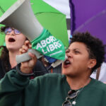 A person holds a megaphone with "Aborto Libre" written on it, passionately speaking during a protest. Surrounding them are others holding colorful umbrellas, suggesting a lively and spirited demonstration.