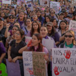 A large group of people, mostly women, gather for a protest. They hold signs in Spanish advocating for women's rights and justice. Many wear purple clothing and bandanas, symbolizing support for the cause. The crowd appears determined and united.