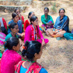 A group of women, dressed in colorful traditional clothing, sit on the ground in a circle outdoors, engaged in conversation or listening intently to a person wearing an Ipas orange cap and a dark t-shirt. The scene appears to be in a rural, wooded area.