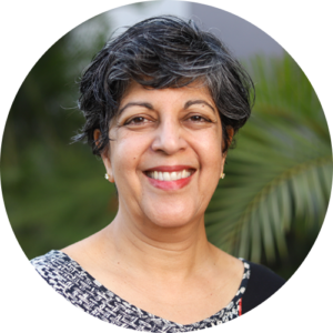Portrait of Dr. Anu Kumar. She has short, gray and black hair is wearing a patterned top and standing in front of greenery. She is smiling and is looking directly at the camera.