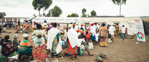 A large group of people, many wearing brightly colored clothing, gather outside a makeshift tent and building in a rural setting amidst the backdrop of a humanitarian crisis. Some are sitting on the rocks while others are standing or walking. There is a sign with the word 