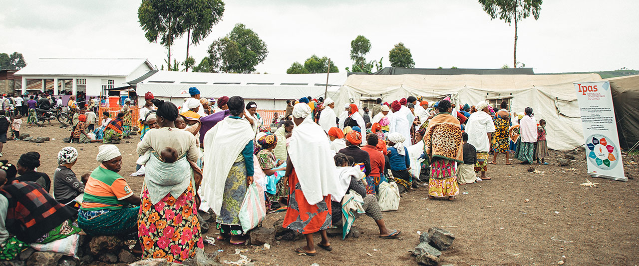 A large group of people, many wearing brightly colored clothing, gather outside a makeshift tent and building in a rural setting amidst the backdrop of a humanitarian crisis. Some are sitting on the rocks while others are standing or walking. There is a sign with the word "Ipas" visible on the right side.
