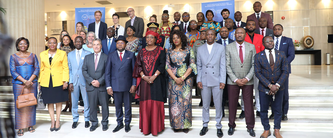 A diverse group of people, including men and women in formal attire, pose for a group photo in a well-lit, modern setting with stairs and banners in the background.