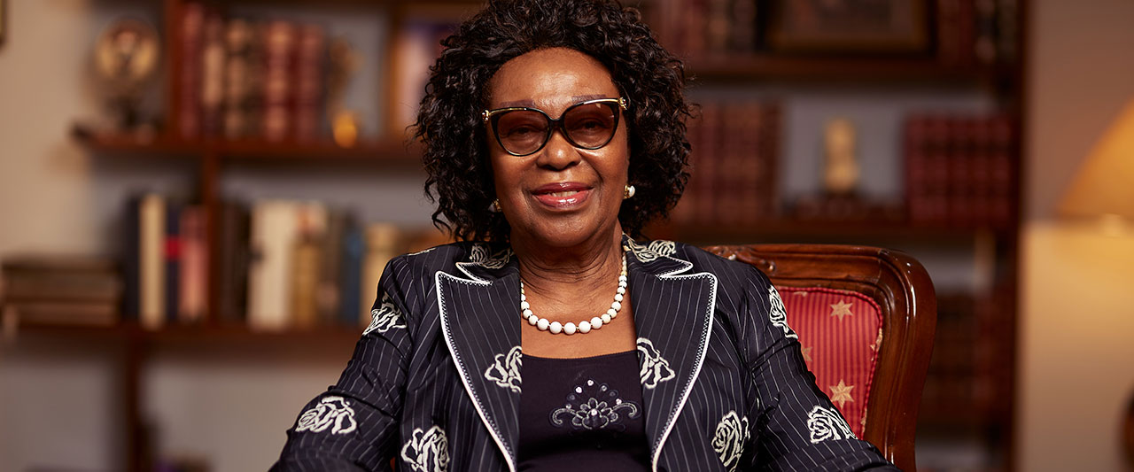 Dr. Eunice Brookman Amissah has curly hair and glasses sits confidently in a library. She wears a pinstriped suit jacket with a floral pattern, a pearl necklace, and rings. Shelves filled with books are visible in the background.