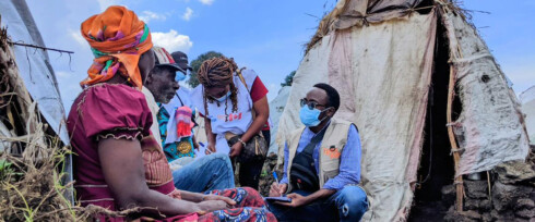 A group of aid workers wearing masks and vests sit outside makeshift tents, discussing something with a local woman in colorful clothing.