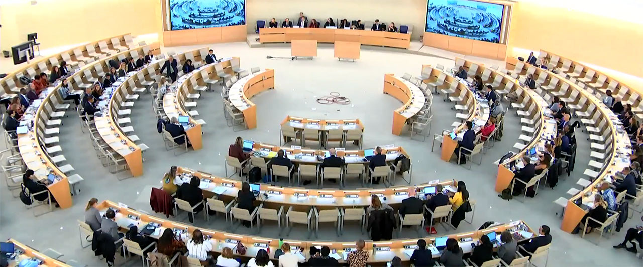The Human Rights Council in a large, semicircular room filled with people seated at rows of desks. Two large screens display an image of the meeting. The main speakers sit at a head table, facing the attendees. Papers and laptops are visible on many of the desks.
