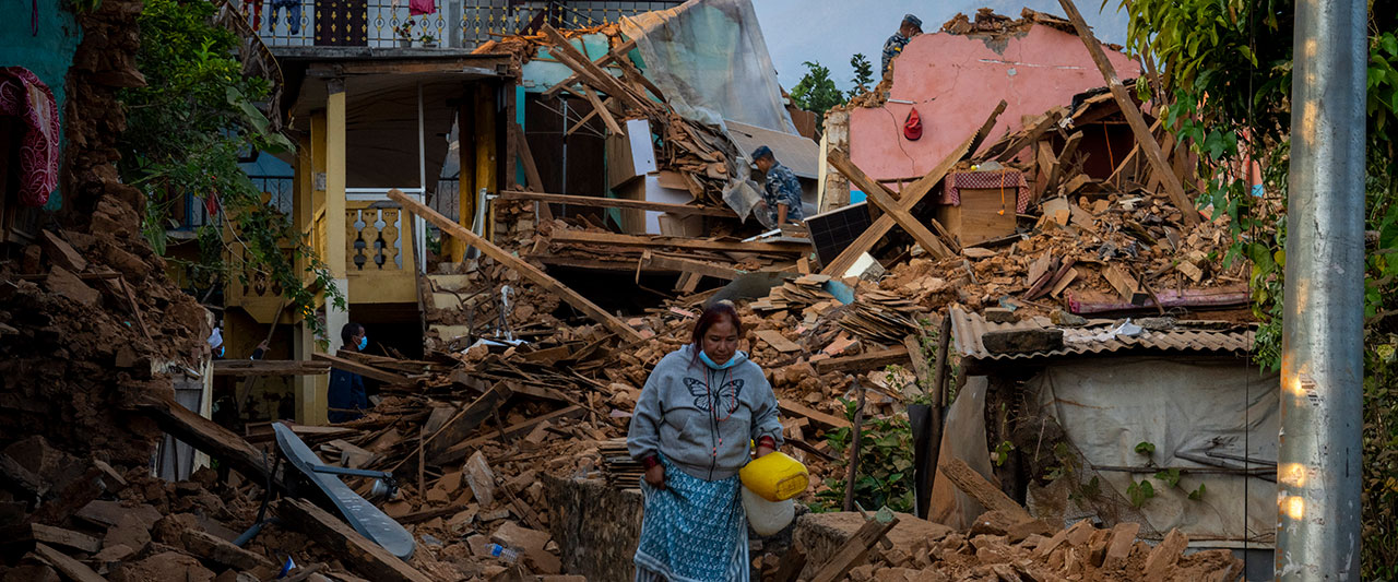 A woman in a gray hoodie stands amidst the rubble of collapsed buildings, carrying a yellow bucket. Surrounding her are remnants of a disaster-struck area, with debris and broken structures. Trees and other damaged houses are visible in the background.
