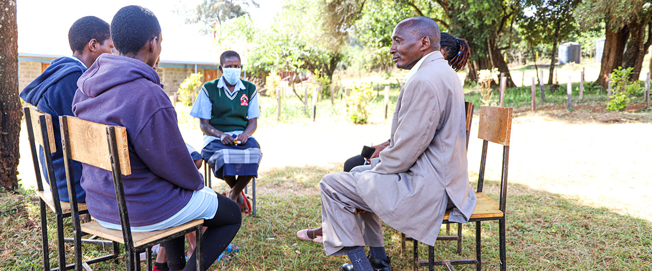 A group of people seated on wooden chairs in a circle outdoors. One person, dressed in a uniform and wearing a face mask, faces the rest who are casually dressed. Amos Simpano, dressed in a light-colored coat is also part of the group. Trees and a building are visible in the background, creating a locally-led atmosphere.