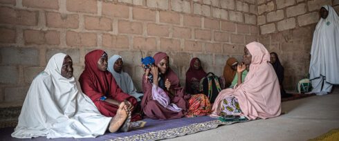 A group of women in colorful attire sit on a carpet in a brick-walled room. Some women wear hijabs in various colors, and they are engaged in conversation, appearing focused and attentive. A larger mat covers part of the floor where they are gathered.