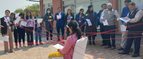 A group of people is participating in an outdoor group activity. One person sits in the center, connected to others by red strings extending outward, each held by a standing participant. They are all holding pieces of paper, engaged in the exercise.