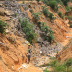 A woman carrying a bucket walks through a rugged, trash-filled ravine surrounded by eroded, reddish-brown earth and sparse green vegetation. The area appears heavily polluted and desolate, an unsettling testament to the effects of climate change.