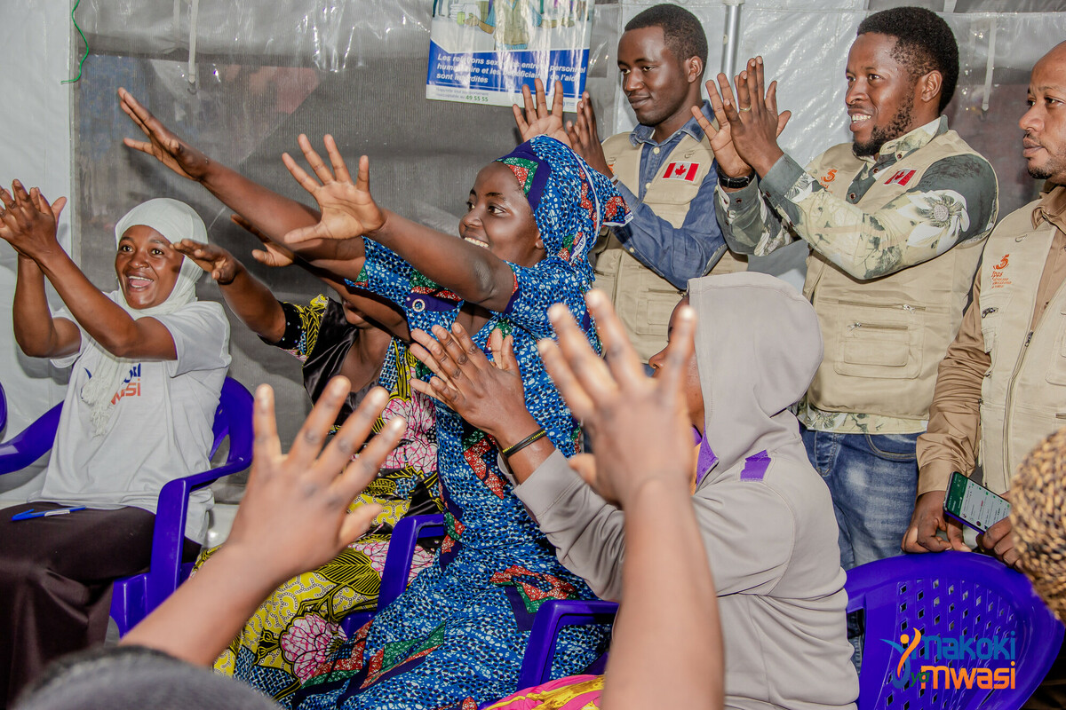 A group of individuals both standing and sitting in chairs, raising their hands in unison and smiling.