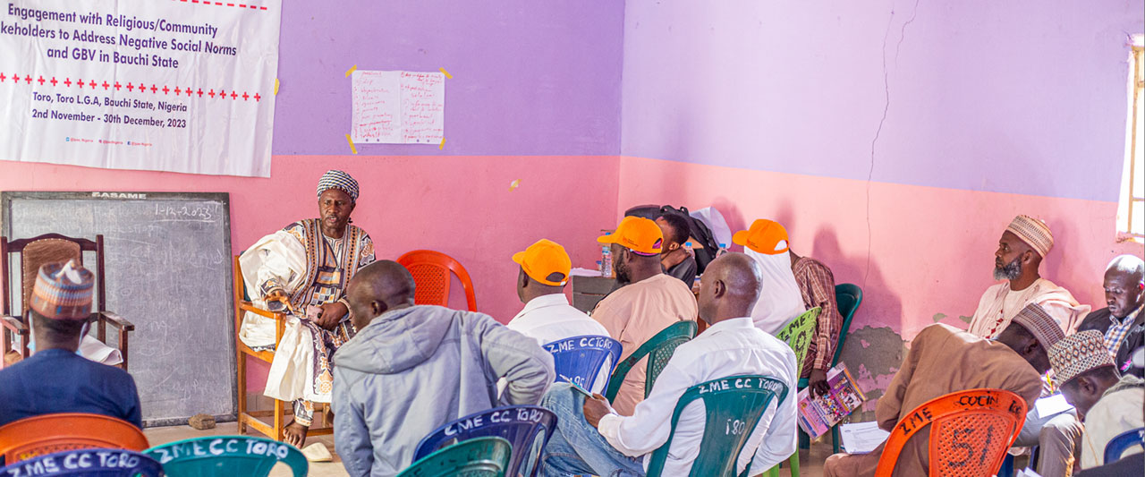 A group of people, some wearing Ipas orange caps, sits in colorful plastic chairs. They are listening attentively to a person dressed in traditional attire seated in front of a banner about gender-based violence and a chalkboard. The room has purple and pink walls.