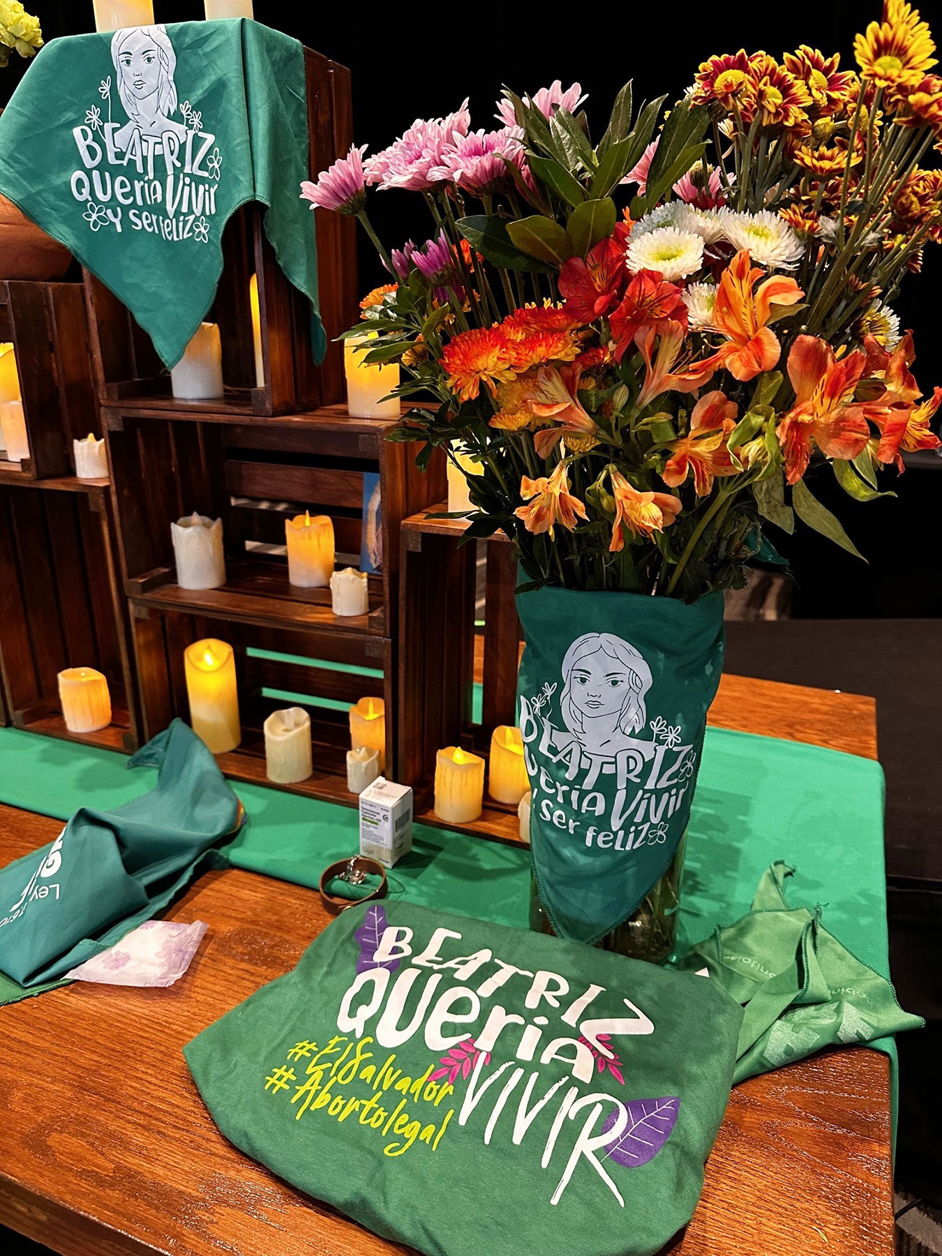 An altar was set up to honor loved ones and to process the suffering caused by lack of abortion access. A display with green bandanas and a green t-shirt, each featuring illustrations and the text "Beatriz Quería Vivir." The table also has a vase of vibrant flowers, a lit candle, and wooden shelves with more candles, creating a memorial-like setting.