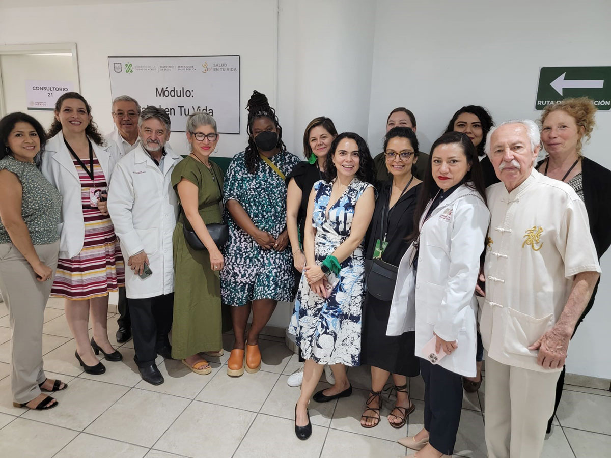 A diverse group of people, including healthcare professionals wearing white coats and individuals in casual attire, smile and pose for a group photo in an indoor setting. A green sign with a white arrow and a poster in Spanish are visible on the wall in the background. A diverse group of people, including healthcare professionals wearing white coats and individuals in casual attire, smile and pose for a group photo in an indoor setting. A green sign with a white arrow and a poster in Spanish are visible on the wall in the background.