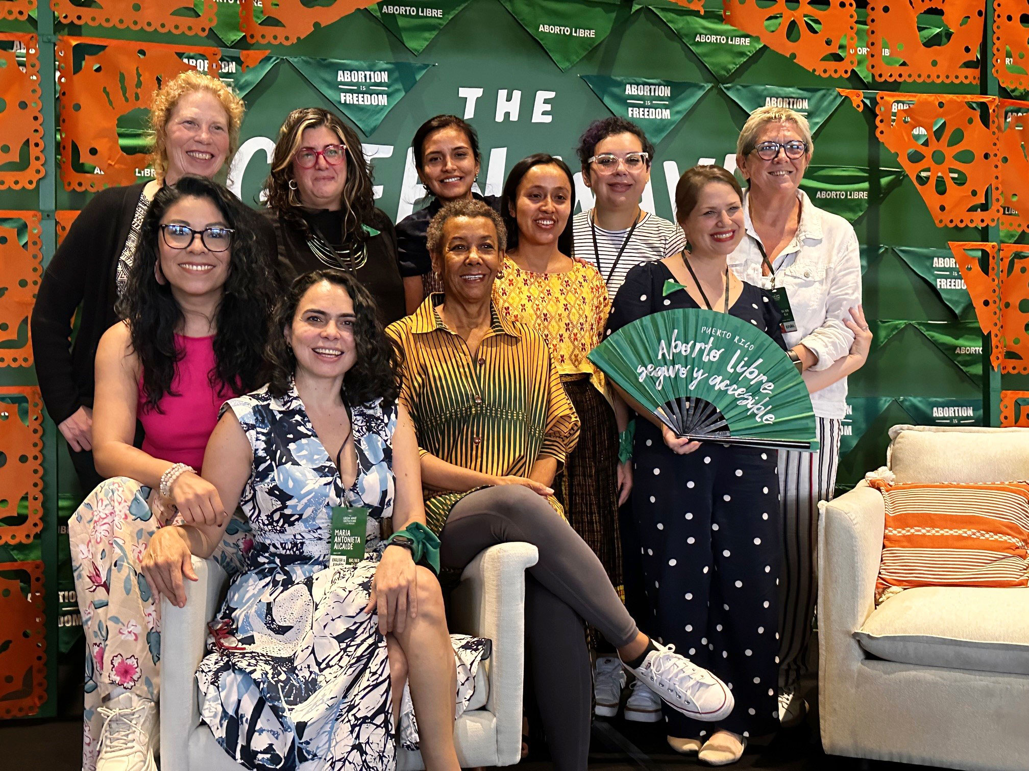 Ipas LAC and US staff A group of ten people smiling and posing together in front of a backdrop with green banners that read "Abortion Freedom" and "Aborto Libre." One person is holding a large green fan with the same text. They are indoors with an orange and green decorated background.