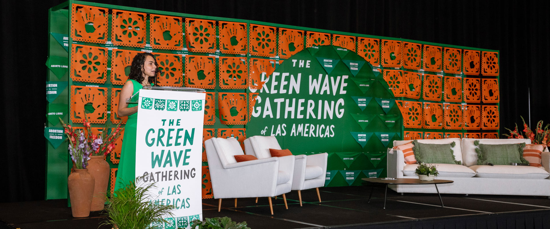 María Antonieta Alcalde Castro, Director, Ipas Latin America and the Caribbean, stands at a podium decorated with "The Green Wave Gathering of Las Americas" text. Beside her are white chairs and a white sofa adorned with green and orange pillows. The backdrop features a vibrant green and orange geometric design, representing the spirit of the Green Wave Gathering.