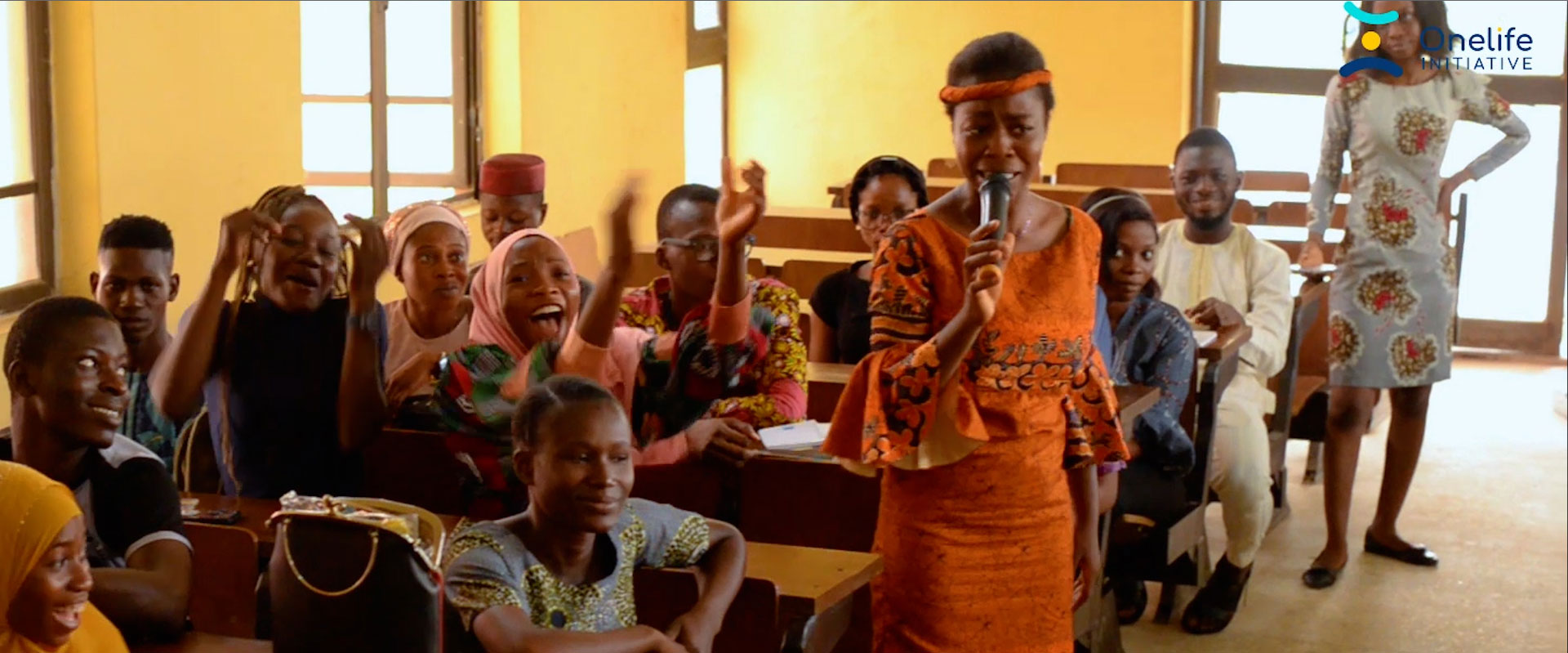 A young woman in an orange dress stands speaking into a microphone in a Nigerian classroom full of smiling students who are raising their hands in joy reacting to her speech