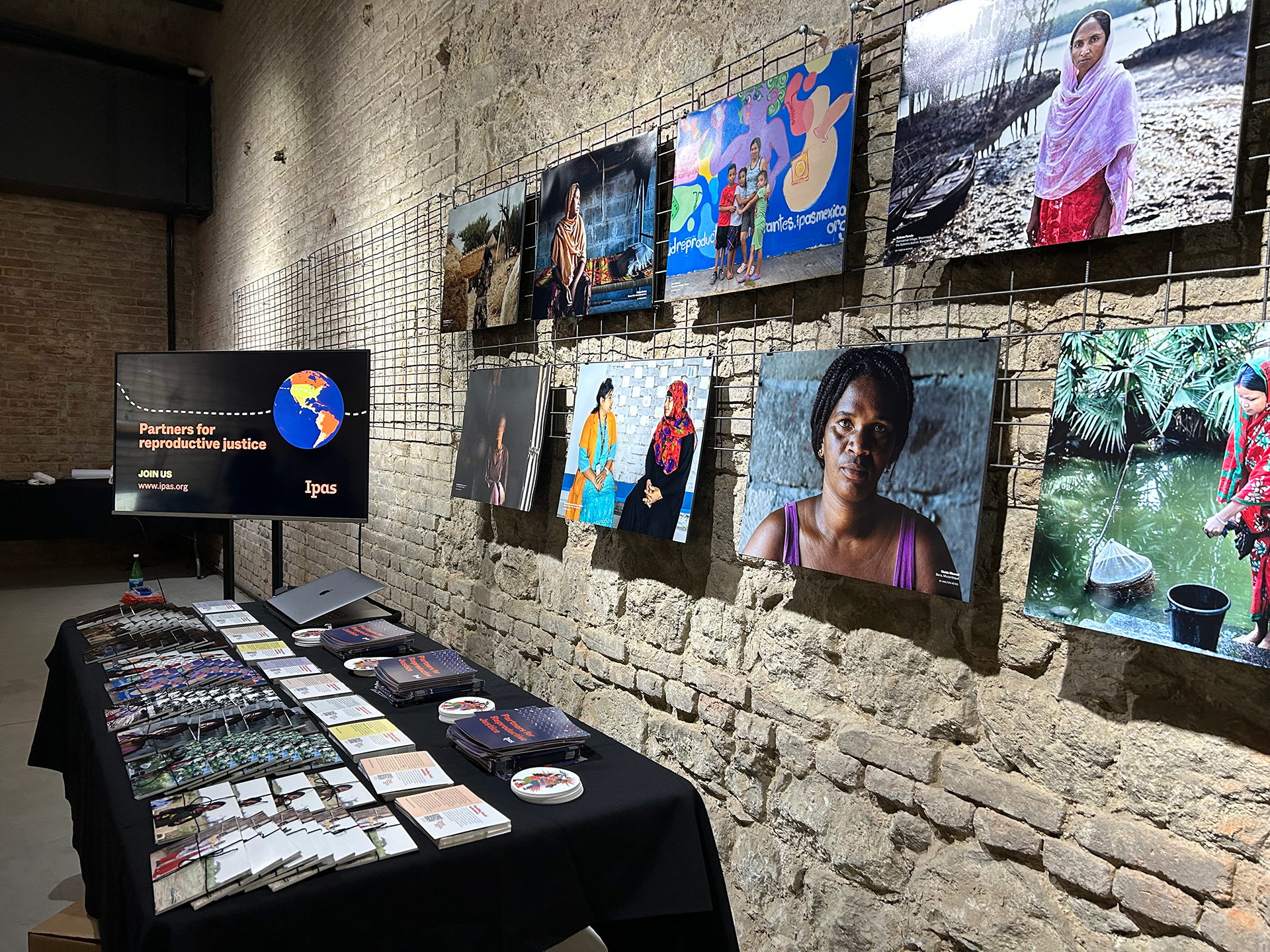 Ipas's photo exhibit at HRFN's Funding Futures Festival A dimly lit gallery showcases a series of photographs of diverse women on a textured brick wall. Below, a table is arranged with pamphlets, booklets, and promotional materials. A screen on the left reads "Partners for reproductive justice" with the Ipas logo.