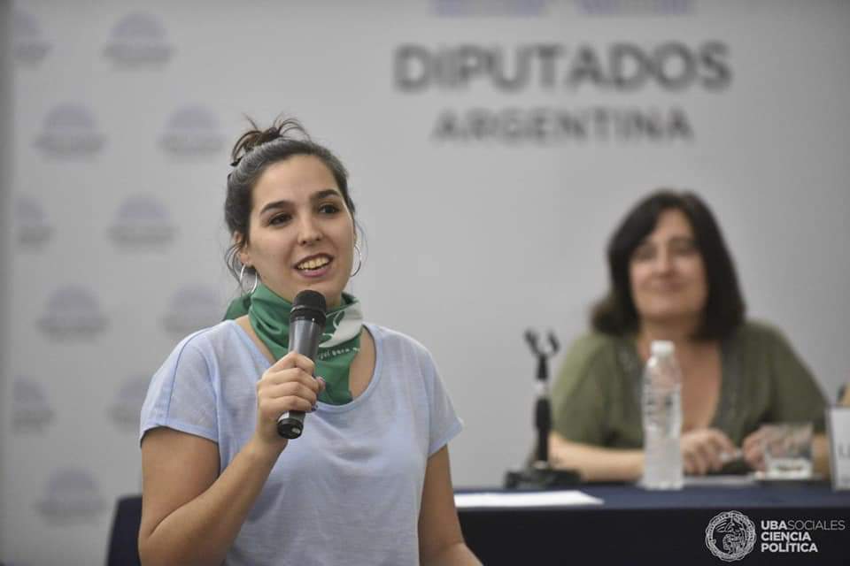 A woman holds a microphone and speaks at an event. She wears a green bandana around her neck and stands in front of a banner that reads, "Diputados Argentina." Another woman sits at a table in the background with a microphone and a water bottle. A woman holds a microphone and speaks at an event. She wears a green bandana around her neck and stands in front of a banner that reads, "Diputados Argentina." Another woman sits at a table in the background with a microphone and a water bottle.