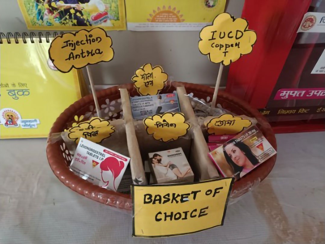 A basket labeled "Basket of Choice" contains various contraceptive options. The basket holds signs for Injection Antira, IUCD copper T, and other contraceptives. Boxes of different contraception methods are organized within the basket. A basket labeled "Basket of Choice" contains various contraceptive options. The basket holds signs for Injection Antira, IUCD copper T, and other contraceptives. Boxes of different contraception methods are organized within the basket.