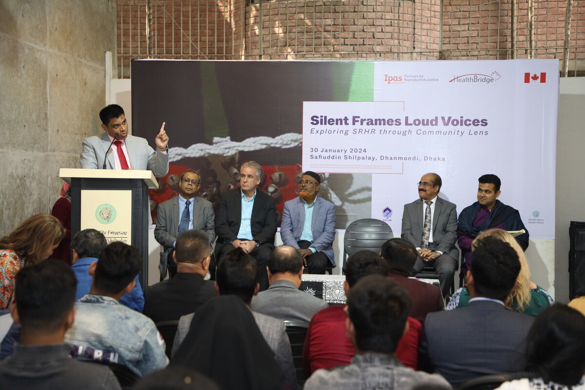 A man in a red tie speaks at a podium in front of an audience. Behind him, a banner reads "Silent Frames Loud Voices: Exploring SRHR through Community Lens," with logos from Ipsos and Global Affairs Canada. Five seated men are on stage, attentively listening. A man in a red tie speaks at a podium in front of an audience. Behind him, a banner reads "Silent Frames Loud Voices: Exploring SRHR through Community Lens," with logos from Ipsos and Global Affairs Canada. Five seated men are on stage, attentively listening.
