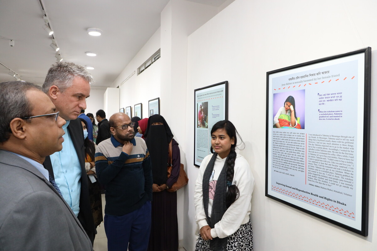 A group of people stand in an art gallery, closely observing framed pieces of text and images displayed on the wall. One woman in the front wears a white sweater and has braided hair. The wall displays detailed artworks or documents with accompanying descriptions. A group of people stand in an art gallery, closely observing framed pieces of text and images displayed on the wall. One woman in the front wears a white sweater and has braided hair. The wall displays detailed artworks or documents with accompanying descriptions.