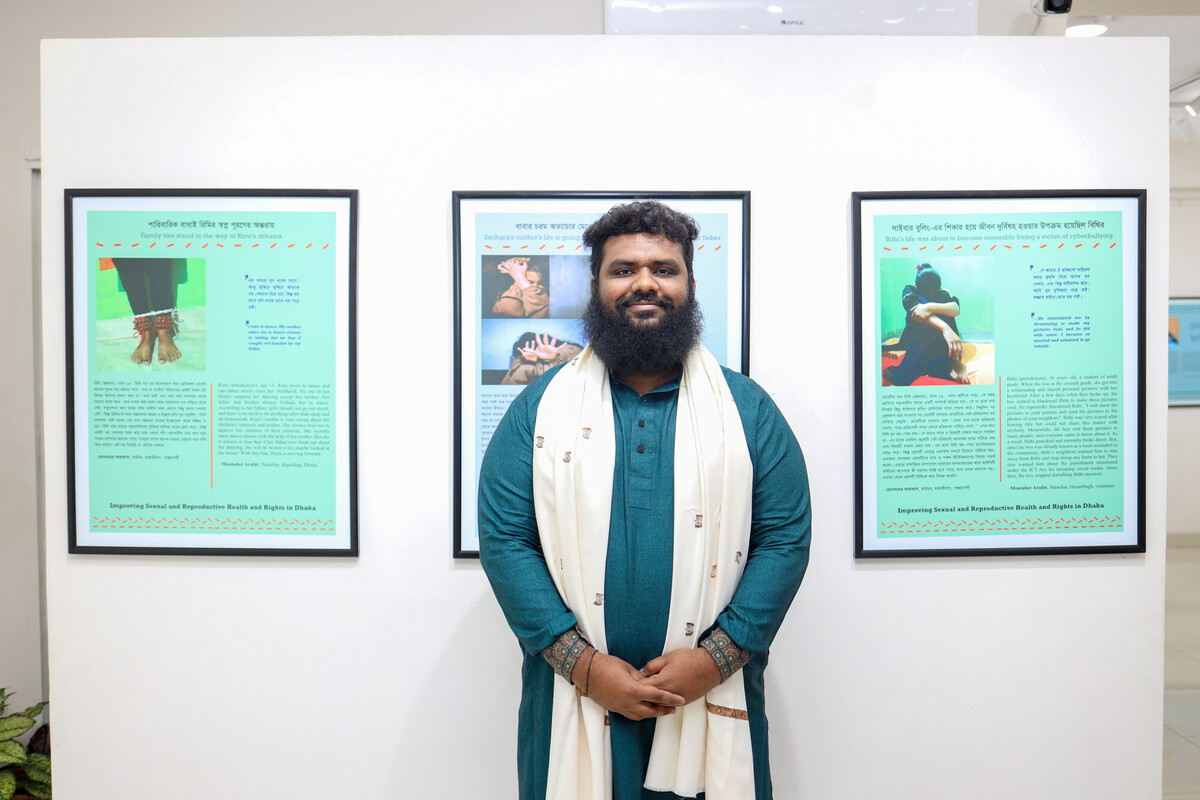 A man with a beard and dark hair, wearing a teal kurta and white shawl, stands in front of a wall displaying three framed informational posters. The posters contain text and images, possibly part of an exhibition. The room appears to be a gallery or display area. A man with a beard and dark hair, wearing a teal kurta and white shawl, stands in front of a wall displaying three framed informational posters. The posters contain text and images, possibly part of an exhibition. The room appears to be a gallery or display area.
