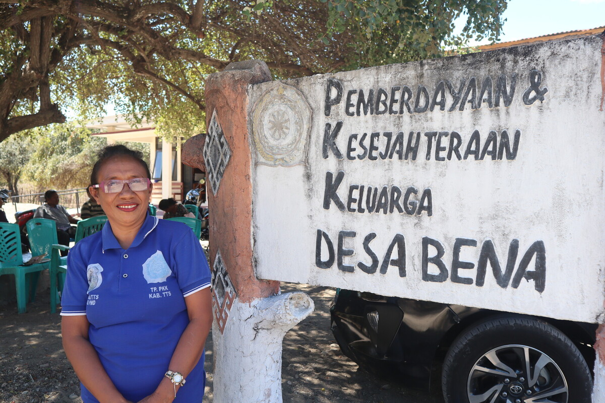 A woman wearing a blue polo shirt and glasses stands smiling beside a large weathered sign that reads, "Pemberdayaan & Kesejahteraan Keluarga Desa Bena." Trees and several people sitting in green chairs are visible in the background. A woman wearing a blue polo shirt and glasses stands smiling beside a large weathered sign that reads, "Pemberdayaan & Kesejahteraan Keluarga Desa Bena." Trees and several people sitting in green chairs are visible in the background.
