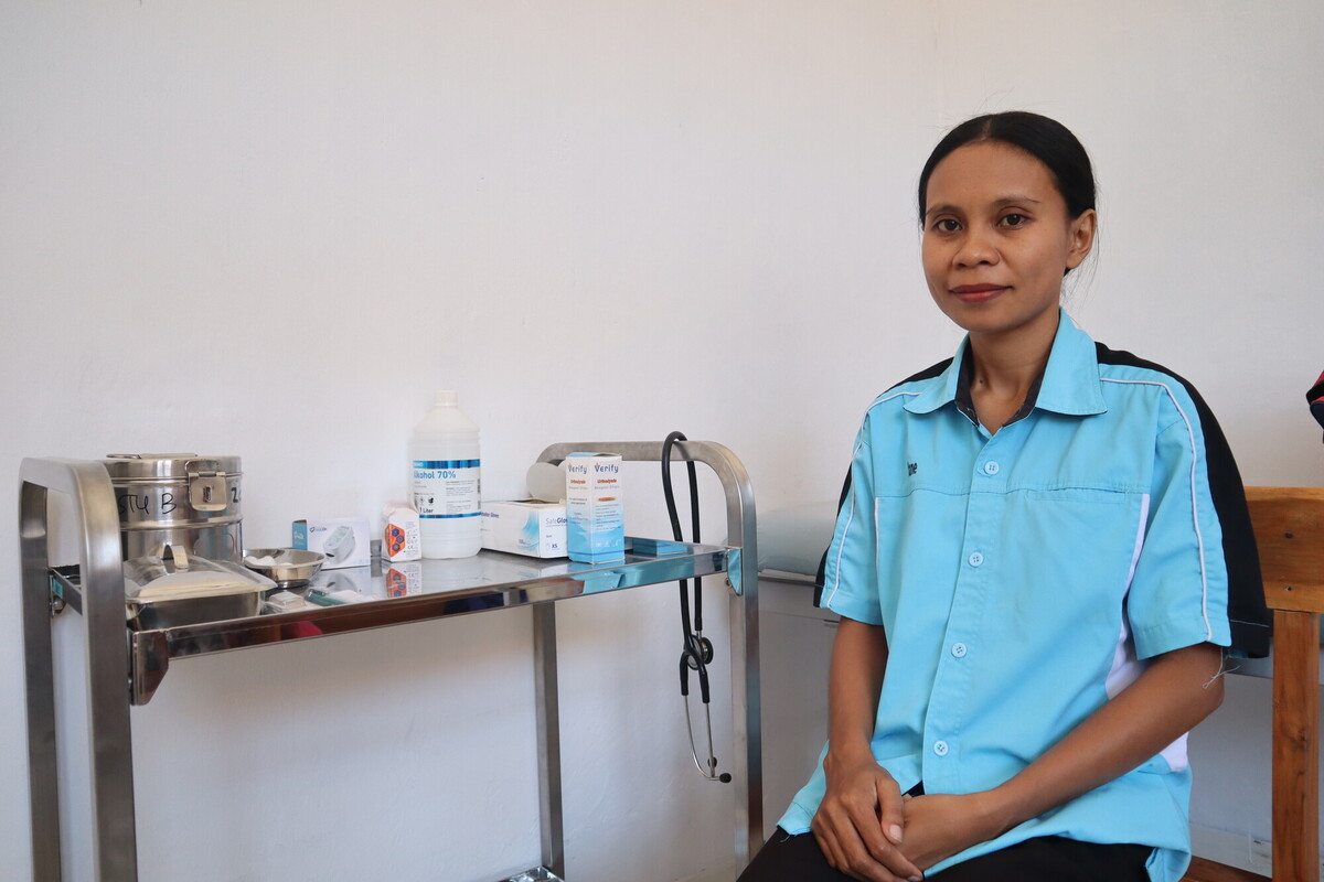 A healthcare worker wearing a light blue uniform sits beside a medical cart containing various supplies, such as a stethoscope, alcohol bottle, and medical boxes, against a plain white wall background. A healthcare worker wearing a light blue uniform sits beside a medical cart containing various supplies, such as a stethoscope, alcohol bottle, and medical boxes, against a plain white wall background.