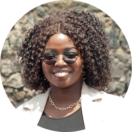 A woman with curly hair and sunglasses smiles against a rocky background. She is wearing a black top, a light-colored blazer, and a gold chain necklace. A woman with curly hair and sunglasses smiles against a rocky background. She is wearing a black top, a light-colored blazer, and a gold chain necklace.