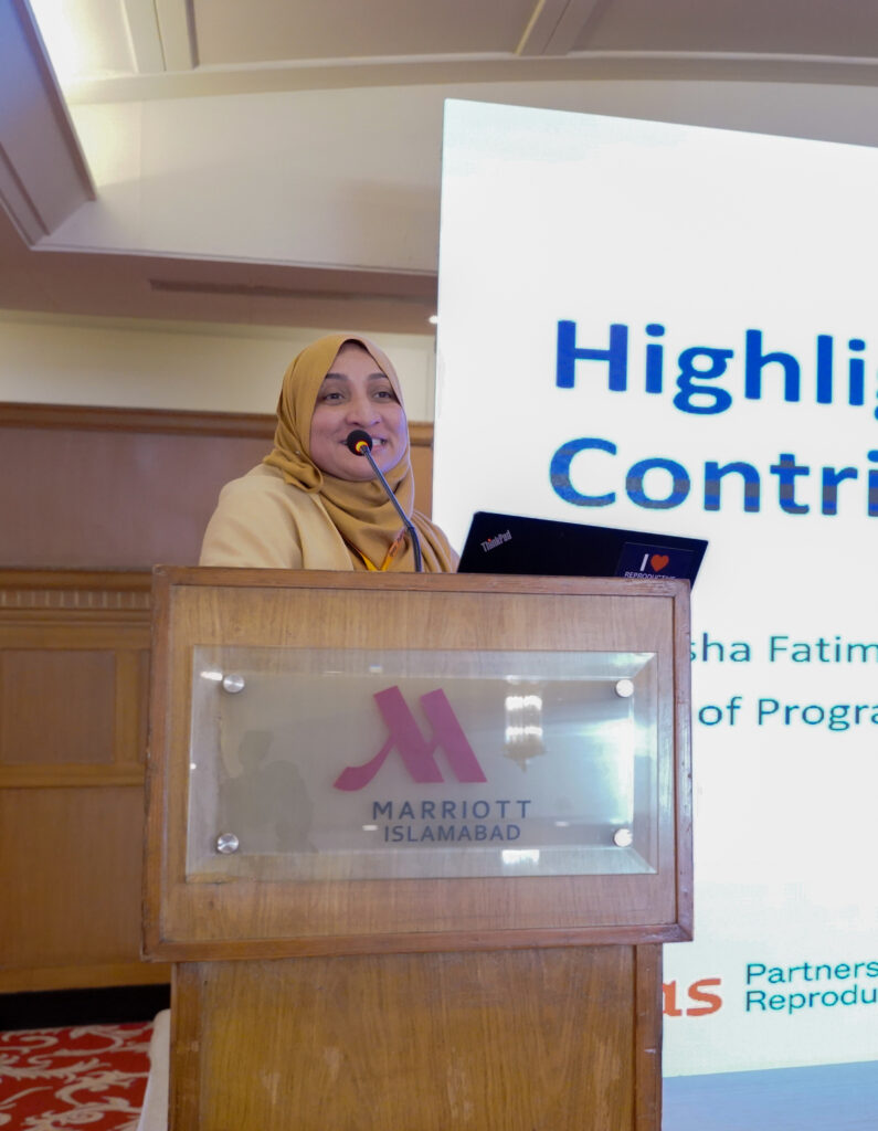 A person wearing a yellow hijab speaks at a podium with a Marriott Islamabad logo. A large screen behind them displays partially visible text in blue. A person wearing a yellow hijab speaks at a podium with a Marriott Islamabad logo. A large screen behind them displays partially visible text in blue.