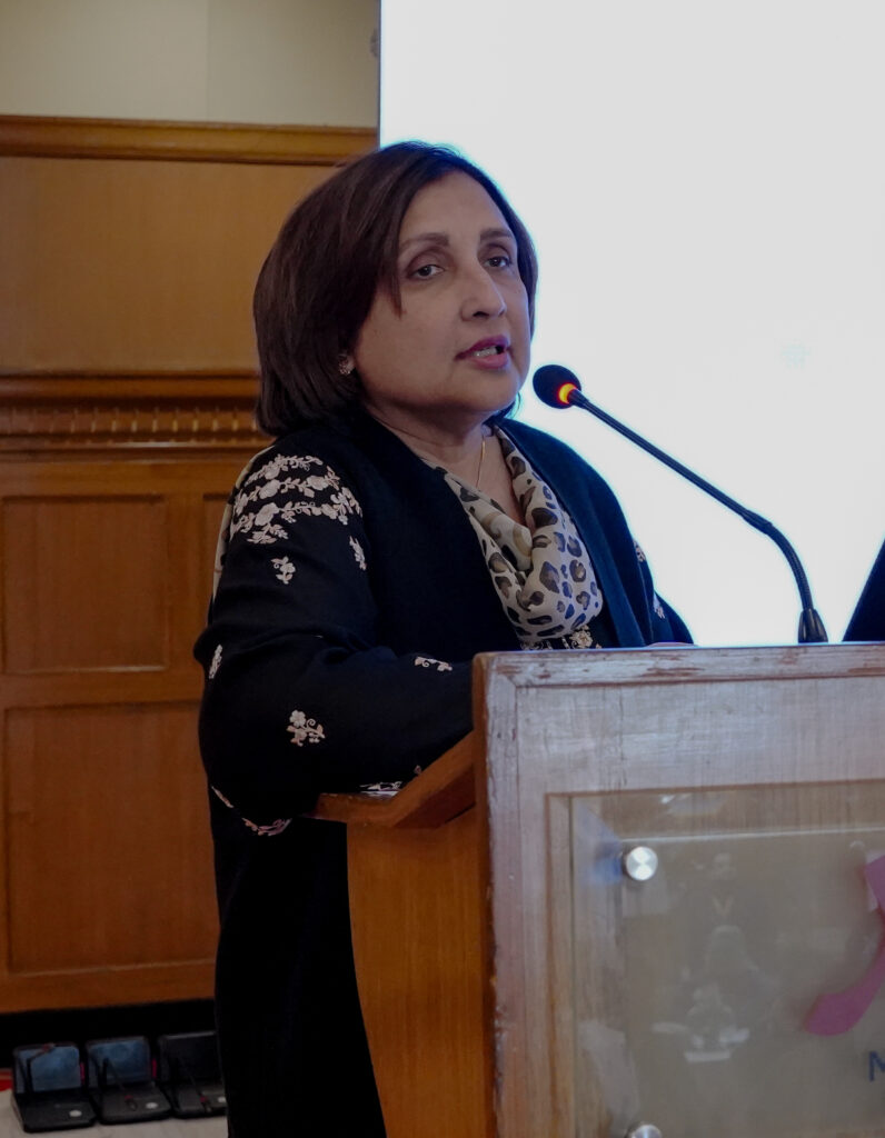 A woman with short dark hair speaks into a microphone at a wooden podium. She is wearing a black and floral outfit. A bright white screen is visible in the background. A woman with short dark hair speaks into a microphone at a wooden podium. She is wearing a black and floral outfit. A bright white screen is visible in the background.