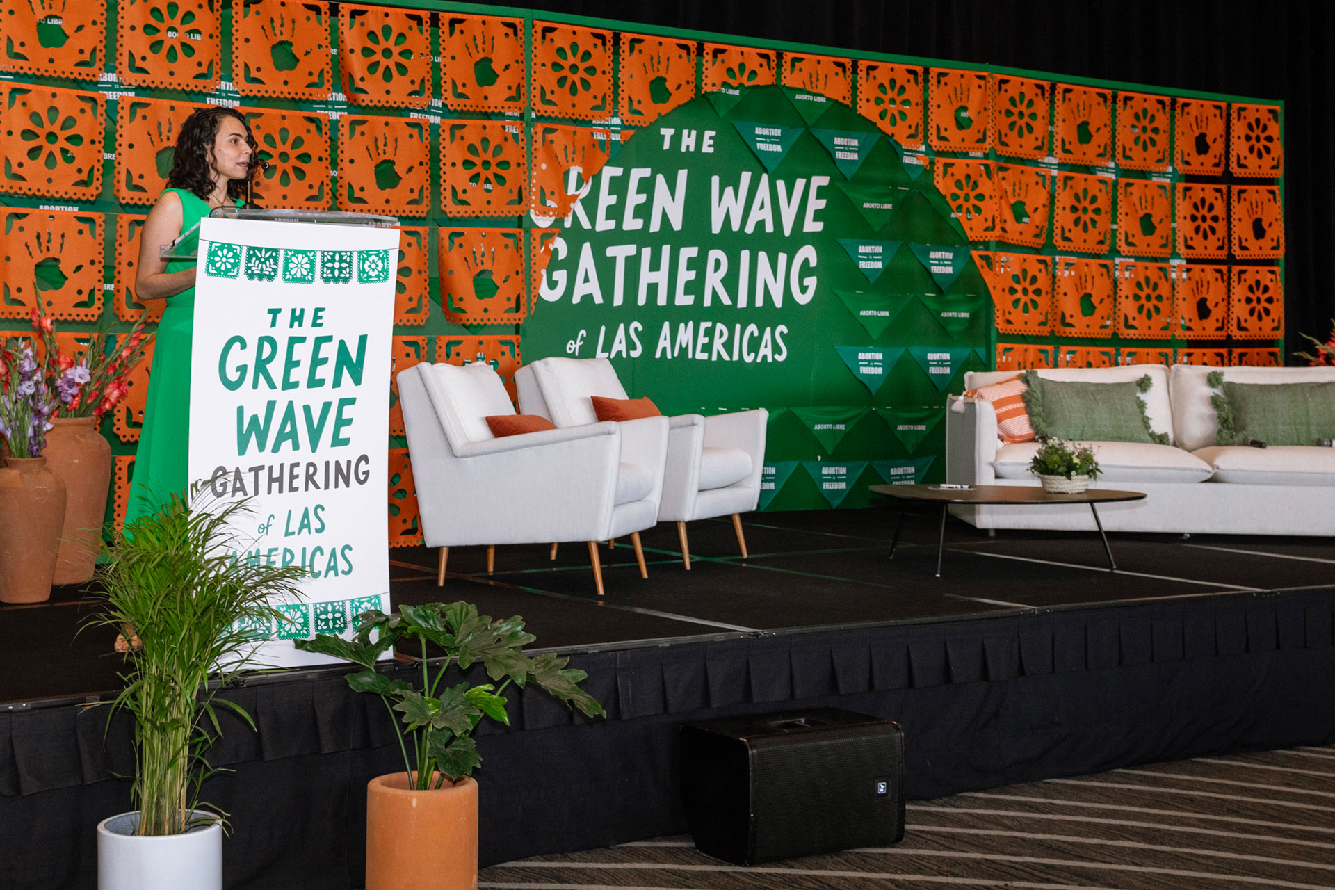 Ipas and partners hosted the second Green Wave Gathering in Mexico City, uniting 150 activists from across the Americas. María Antonieta Alcalde Castro, Director, Ipas Latin America and the Caribbean, stands at a podium decorated with "The Green Wave Gathering of Las Americas" text. Beside her are white chairs and a white sofa adorned with green and orange pillows. The backdrop features a vibrant green and orange geometric design, representing the spirit of the Green Wave Gathering.