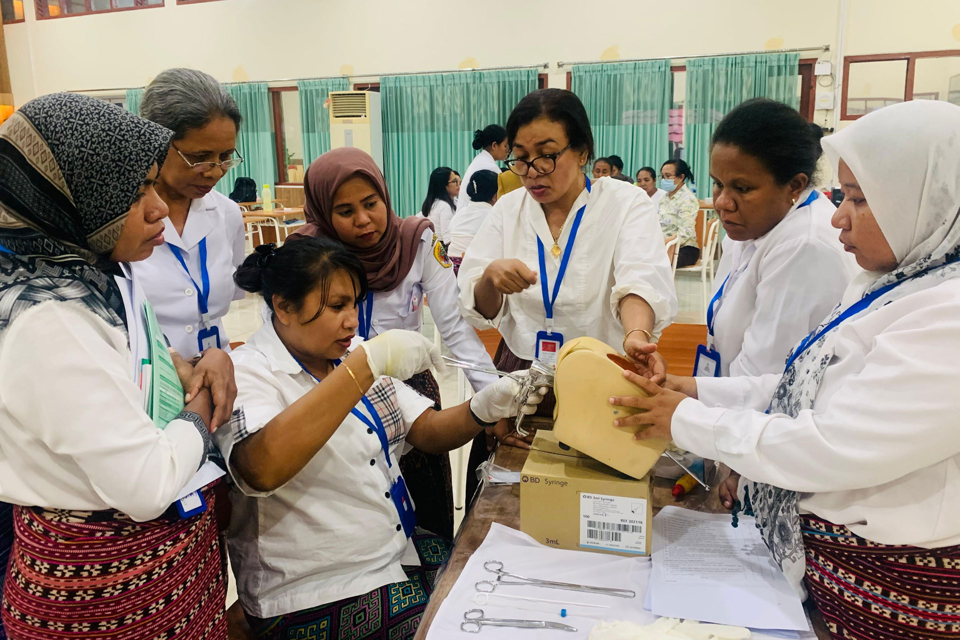 Midwives in NTT participated in the family planning training A group of women in white uniforms, some wearing headscarves, gather around a table. One woman demonstrates a medical procedure on a training model while others watch attentively. Surgical tools and a manual are visible on the table.