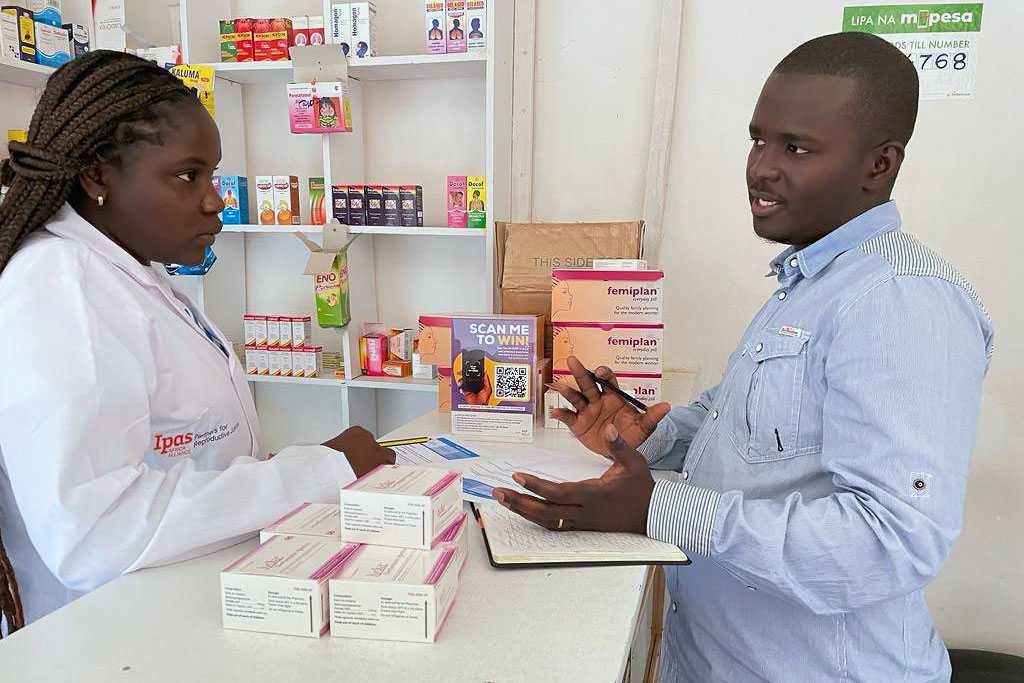 Ipas partner pharmacists for medical abortion self-use and post abortion contraception service delivery. A pharmacist in a white coat discusses medication details with a man holding a pen and notebook. They are surrounded by medicine boxes and health products on shelves in a pharmacy setting.