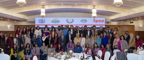 A large group of people, including men and women, pose together indoors in front of a display with the logos of various organizations. The setting includes tables with floral decorations and elegant lighting fixtures.