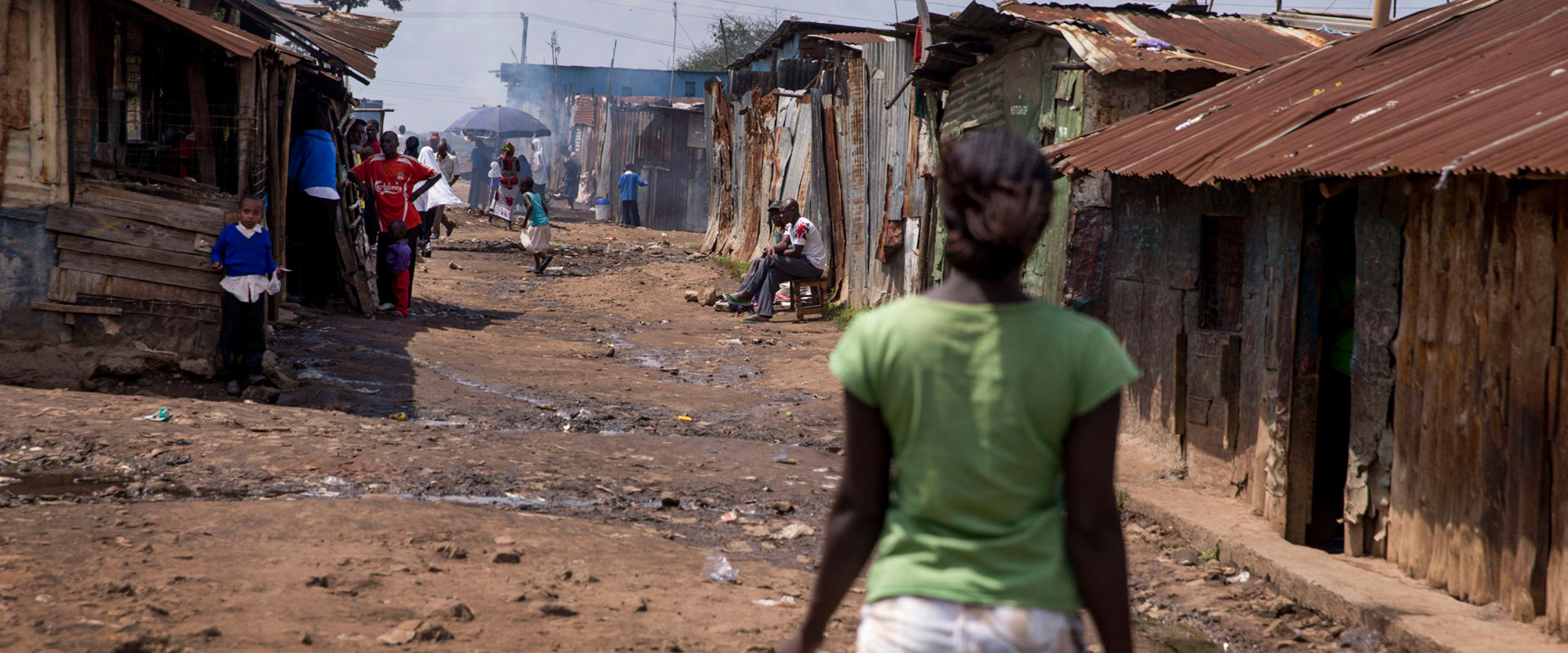 A dirt road lined with rustic houses. People, including children, walk along the road. The area appears to be a densely populated, informal settlement. A person in a green shirt is in the foreground walking away.