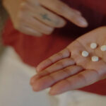 A person wearing a red shirt holds four small white pills in their open palm. Their other hand is raised with one finger pointing towards the pills. Subtle tattoos are visible on both wrists.