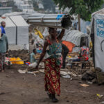 A woman in a colorful dress and headscarf carries a metal sheet on her head through a an camp for Internally Displaced People (IDP) with tents and shelters around. Other people are visible in the background, some seated, amidst a rugged, rocky terrain.