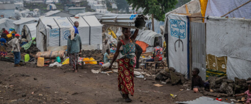 A woman in a colorful dress and headscarf carries a metal sheet on her head through a an camp for Internally Displaced People (IDP) with tents and shelters around. Other people are visible in the background, some seated, amidst a rugged, rocky terrain.