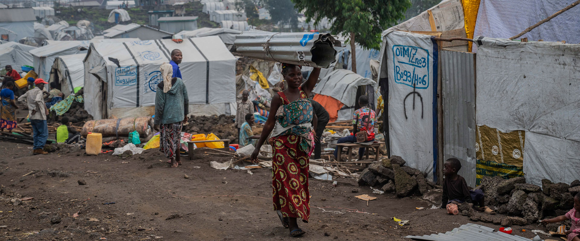 A woman in a colorful dress and headscarf carries a metal sheet on her head through a an camp for Internally Displaced People (IDP) with tents and shelters around. Other people are visible in the background, some seated, amidst a rugged, rocky terrain.