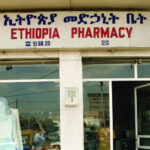 Front view of a pharmacy with a sign in Amharic and English reading "Ethiopia Pharmacy." A mannequin is visible in the window next to the entrance. The building exterior is beige with a purple section on the side.