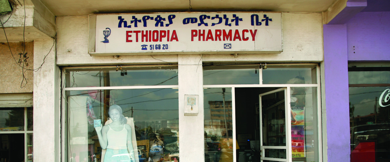 Front view of a pharmacy with a sign in Amharic and English reading "Ethiopia Pharmacy." A mannequin is visible in the window next to the entrance. The building exterior is beige with a purple section on the side.