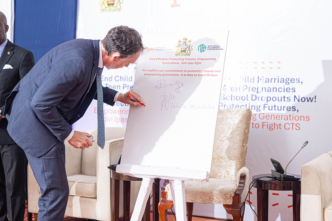 The Ambassador of Iceland in Malawi, signing commitment for support of CTS. A man in a suit signs a large board at an event focused on ending child and teen pregnancies. The board has logos and text supporting the cause. There's a man in the background and a chair and speaker to the side.