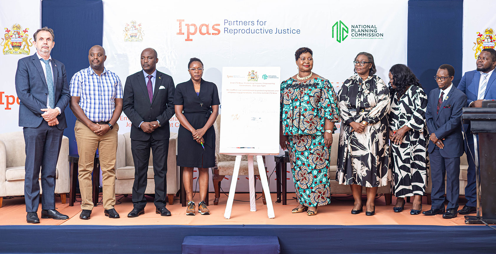 Delegates who signed the commitment on supporting CTS A group of nine people stands on a stage in front of a backdrop featuring "Ipas" and "National Planning Commission" logos. They are posing next to a large signed document, dressed in formal and business attire.