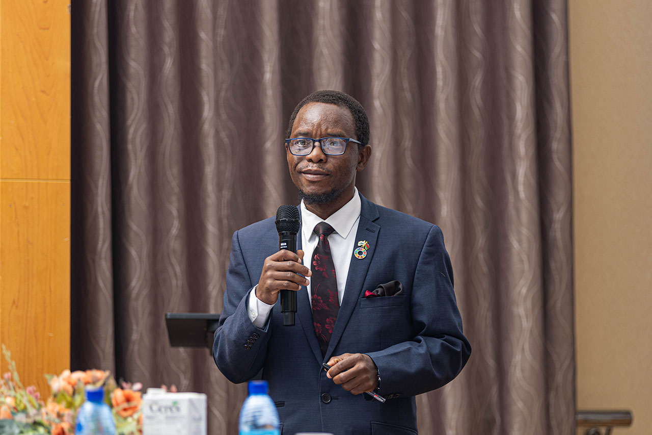 Dr. Andrew Jamali from the National planning commission, making a CTS Malawi case presentation A man in a suit and tie holds a microphone, standing in front of a curtain. He wears glasses and a pin on his lapel. A table with a water bottle and flowers is in the foreground.