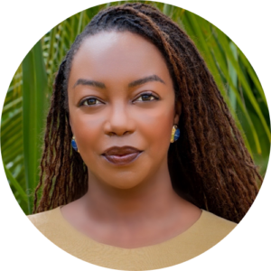 A woman with long, brown, braided hair and blue earrings, wearing a beige top, stands outdoors in front of green palm leaves, smiling softly at the camera.