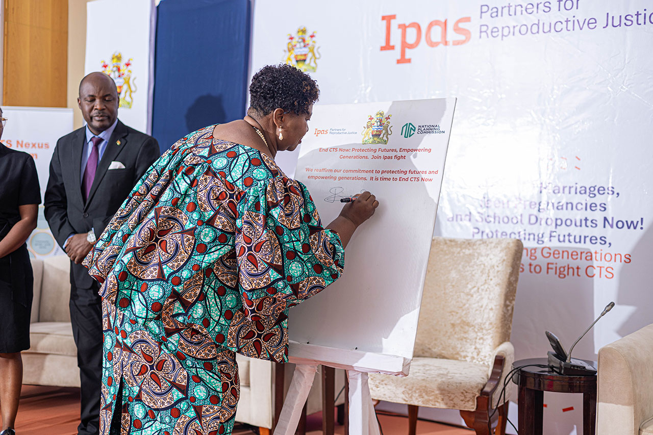 Hon. Minister of Gender, Jean Sendeza signing the commitment of CTS support. A person in patterned attire signs a board during an event. Two others stand nearby, one of whom is wearing a suit. The backdrop displays logos and the text "Ipas Partners for Reproductive Justice." The setting appears formal and focused on activism.