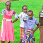 Six smiling girls stand on grass, each holding up a cup of yogurt. Wearing brightly colored dresses, they appear joyful and proud—celebrating support from the Collaborative Fund in raising awareness about period poverty outdoors together.